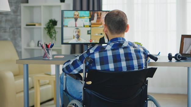 Freelancer in wheelchair waving during a business video call while working from home office.