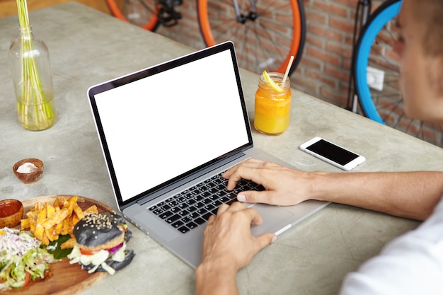 Freelancer in white t-shirt working remotely using laptop computer during lunch, sitting at cafe table with burger