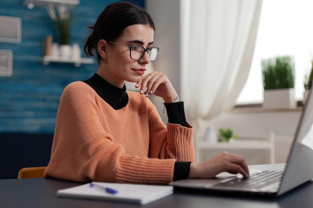 Freelancer with glasses casually typing on laptop keyboard. Student with notebook studying remote smiling at computer screen. Programmer sitting at home desk sending email and browsing the internet.