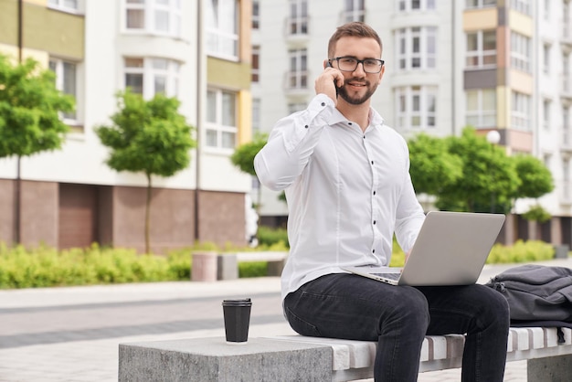 Freelancer with laptop sitting on bench talking by phone
