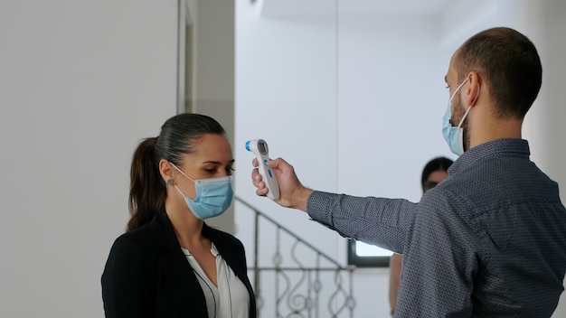 보호용 안면 마스크를 쓴 프리랜서는 동료들이 사무실에 들어가기 전에 온도계로 온도를 측정합니다. 코로나19 감염 예방을 위한 사회적 거리두기 실천하는 동료들