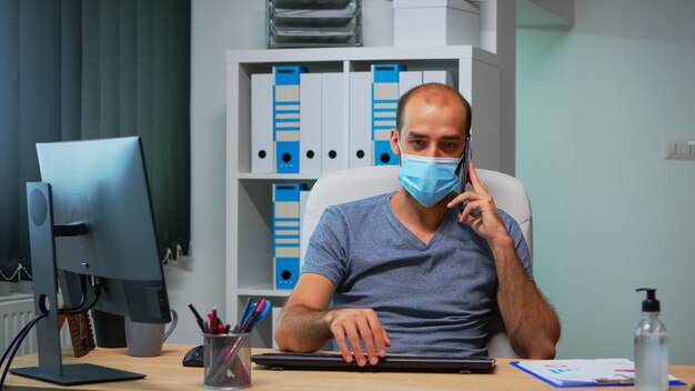 Freelancer with protection mask talking on mobile phone with partners, sitting on desk in office room during pandemic. Freelancer working in new normal office chatting writing speaking on smartphone