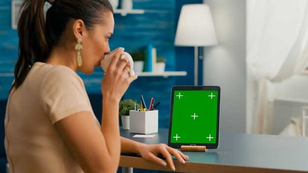 Фрилансер женщина, держащая чашку кофе, смотрящую на планшетный компьютер с макетом зеленого экрана цветности ключа, сидящего на столе стола. Кавказская женщина просматривает изолированное устройство из комнаты домашнего офиса