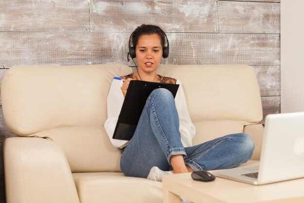 Freelancer woman wearing headphones and taking notes on clipboard during job video call.