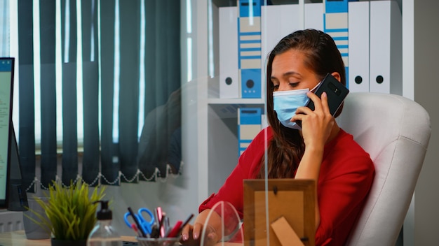 Фрилансер, работающий и разговаривающий по телефону, сидит на рабочем месте в защитной маске во время пандемии коронавируса. Женщина разговаривает с командой удаленно, говоря на смартфоне перед компьютером