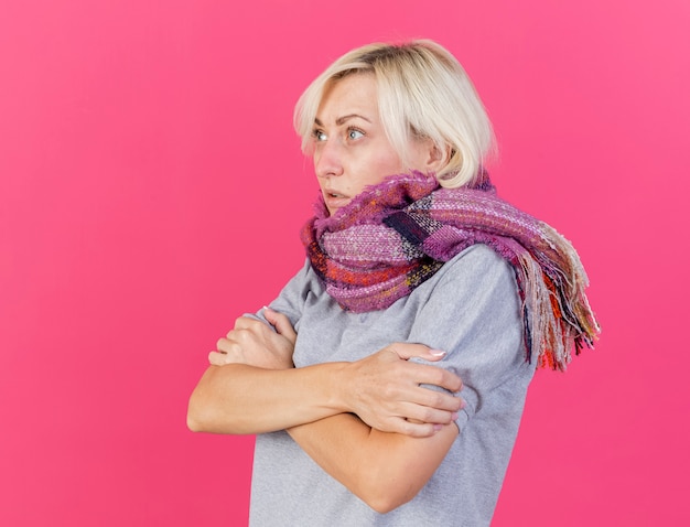 Freezing young blonde ill slavic woman wearing scarf holds arms looking at side on pink