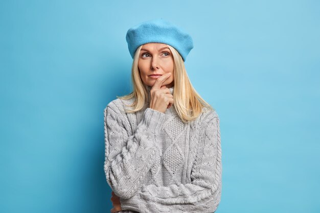 French style. Thoughtful dreamy blonde middle aged European woman thinks about good future holds chin has serious expression dressed in stylish beret and knitted warm sweater for cold weather