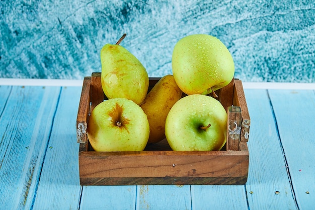 Свежие яблоки и груши в деревянном ящике на синей поверхности.