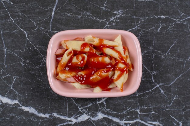 Fresh boiled penne pasta with ketchup in pink bowl.