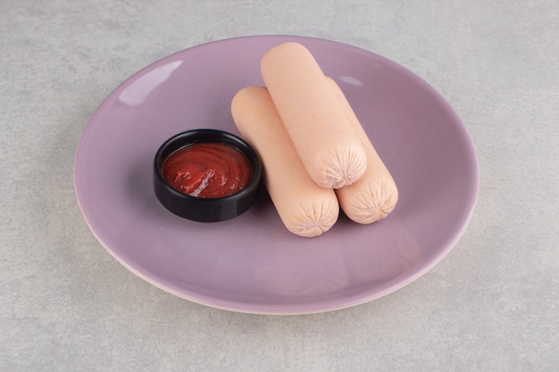 Свежие вареные сосиски и кетчуп на фиолетовой тарелке.