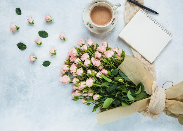 Fresh bouquet of pink roses with cup of coffee; spiral notepad and pen on textured background