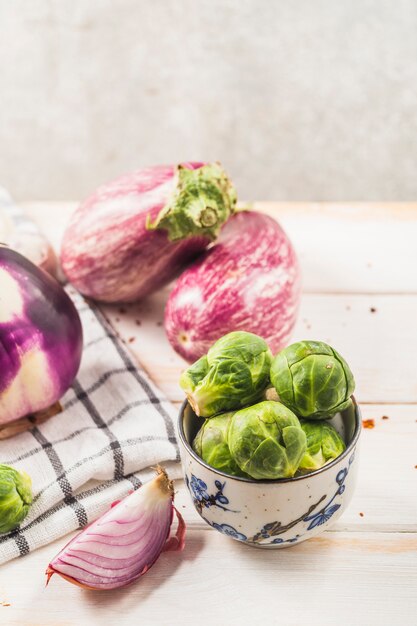 Fresh brussels sprouts in bowl near eggplants; onion and textile on wooden table