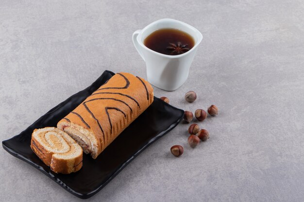 Fresh cake roll with cup of tea on black plate over grey background. 