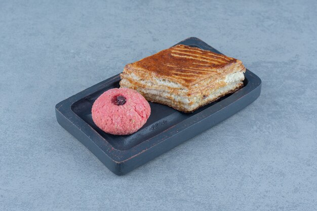 Fresh cake slice with pink cookie on wooden plate.