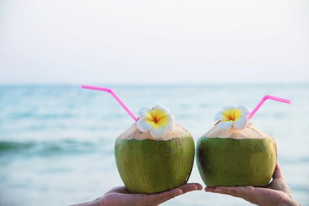 Fresh coconut in couple hands with plumeria decorated on beach with sea wave - honeymoon couple tourist with fresh fruit and sea sand sun vacation concept