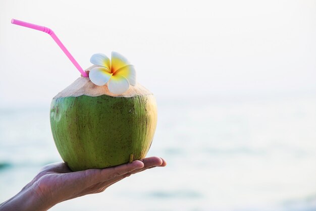Fresh coconut in hand with plumeria decorated on beach with sea wave - tourist with fresh fruit and sea sand sun vacation concept