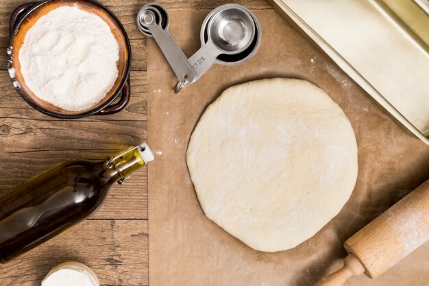 Fresh dough ready for baking on parchment paper with measuring spoons; flour; oil and rolling pin over the wooden desk