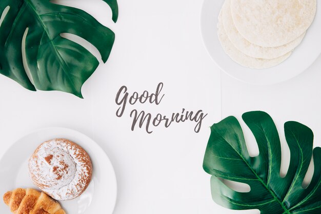 Free Photo fresh flour tortillas; baked bun; croissant breakfast with good morning text on paper and green monster leaves on white background