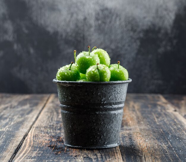Fresh green plums in a mini bucket side view on wooden and misty wall