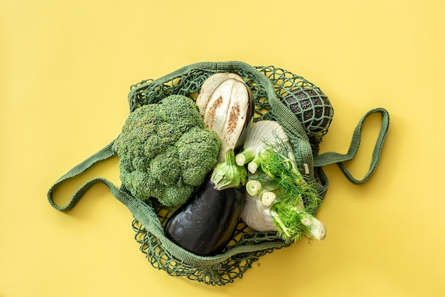 Fresh green vegetables in a green string bag on a yellow background flat lay