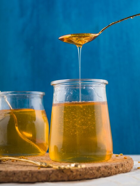 Fresh honey dripping in glass pot on cork coasters against blue background