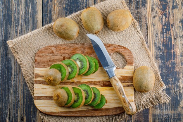 Свежий киви с разделочной доской, ножом на деревянном фоне и кусок мешка, плоская планировка.