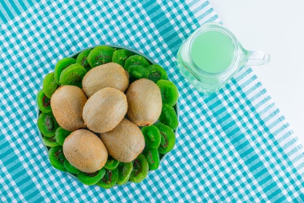 Fresh kiwi with dried kiwi, drink in a plate on picnic cloth and white background, flat lay.