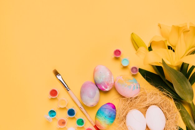 Fresh lily flower with colorful easter eggs; paintbrush and watercolor on yellow background