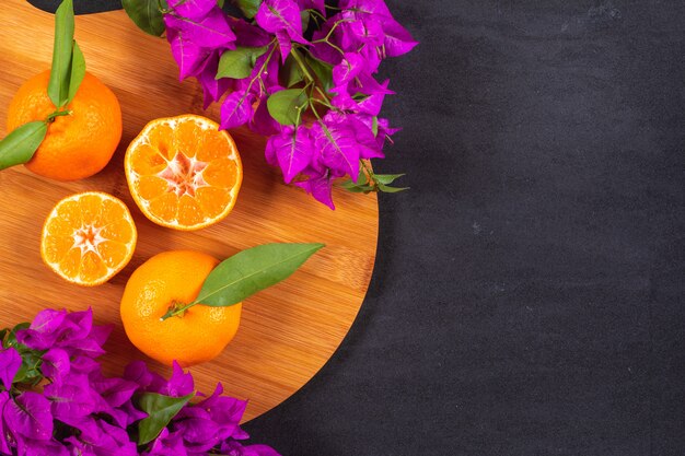 Fresh mandarins on wood cutting board with purple flowers on black surface with copy space