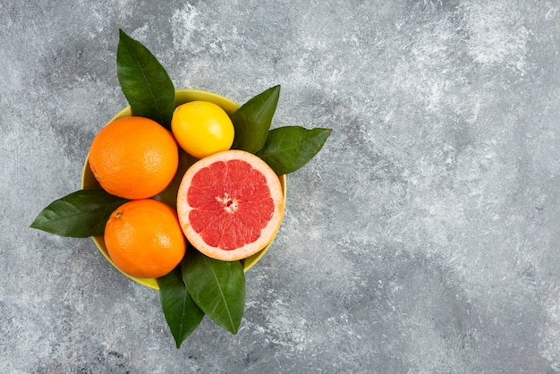 Fresh organic citrus fruits in bowl with leaves over grey table.