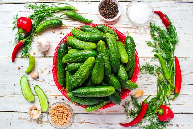 Fresh organic cucumbers in red basket on white wooden table with green and red and chili peppers, fennel, salt, black peppercorns, garlic, pea, close up, healthy concept, top view, flat lay