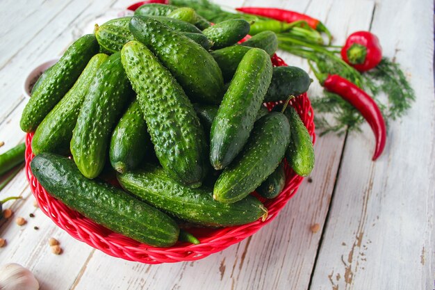 Fresh organic cucumbers in red basket on white wooden table with green and red and chili peppers, fennel, salt, black peppercorns, garlic, pea, close up, healthy concept, top view, flat lay