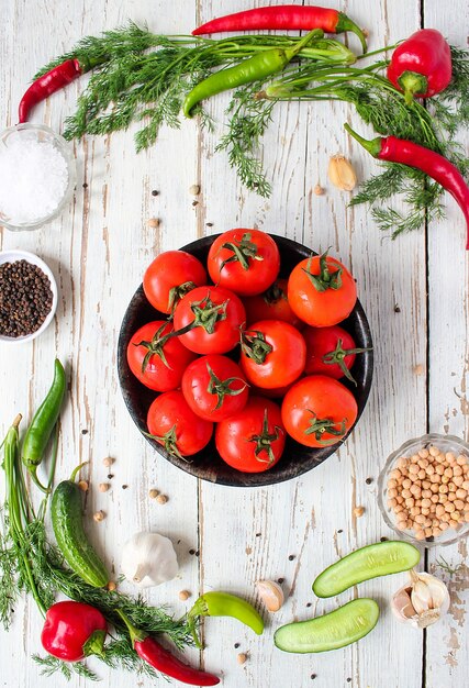 Fresh organic red tomatoes in black plate on white wooden table with green and red and chili peppers, green peppers, black peppercorns, salt, close up, healthy concept