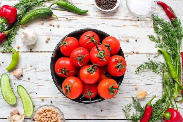 Fresh organic red tomatoes in black plate on white wooden table with green and red and chili peppers, green peppers, black peppercorns, salt, close up, healthy concept