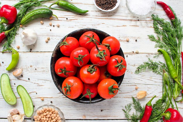 Fresh organic red tomatoes in black plate on white wooden table with green and red and chili peppers, green peppers, black peppercorns, salt, close up, healthy concept