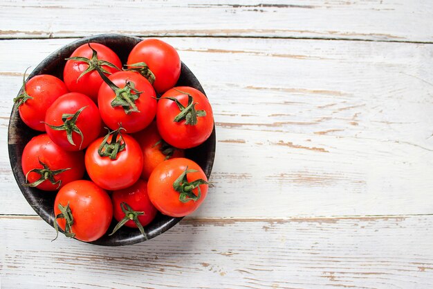 Fresh organic red tomatoes in black plate on white wooden table with green and red and chili peppers, green peppers, black peppercorns, salt, close up, healthy concept