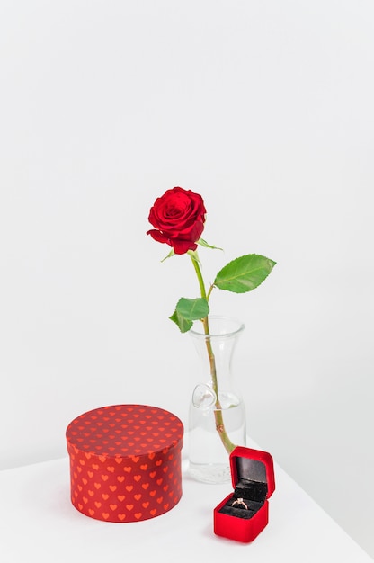 Fresh red rose in vase near present and jewellery box with ring on table