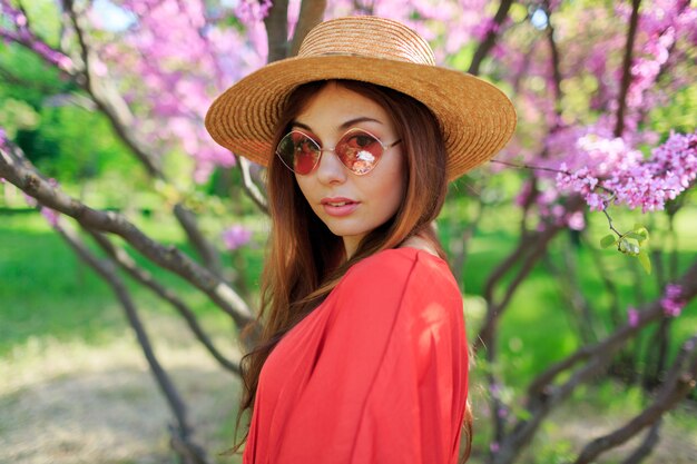 Fresh spring portrait of cute smiling woman in stylish coral dress, in straw hat enjoying sunny day