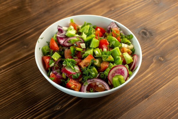 Салат из свежих овощей, включая нарезанные огурцы, красный помидор, лук и другие вещи внутри белой тарелки на деревянной деревенской поверхности