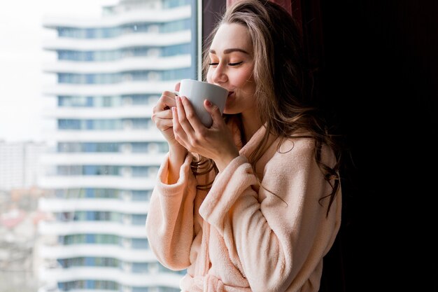 ピンクの柔らかいバスローブで新鮮な若い女性は、窓の外を見てお茶を飲みます。