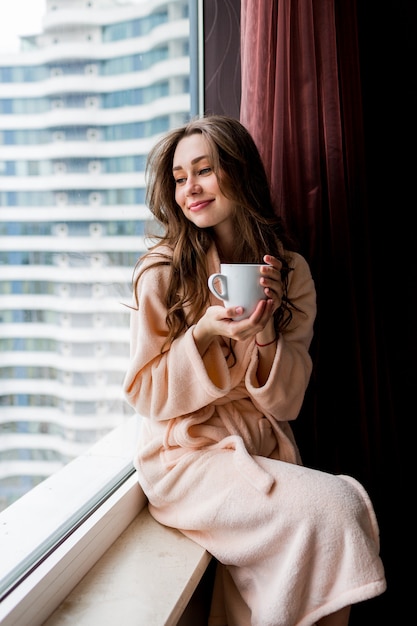 Свежая молодая женщина в розовом нежном халате пьет чай, глядя в окно.