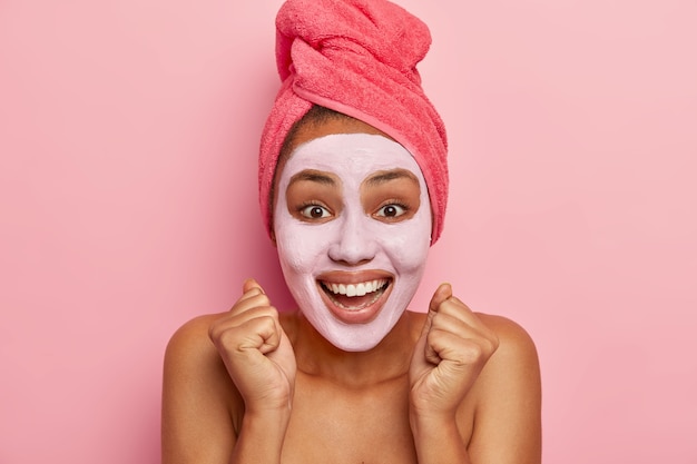 Freshness, shower, self care concept. Overjoyed ethnic lady raises clenched fists, smiles positively, makes beauty treatments after bath, face covered with clay moisturising mask, naked healthy body