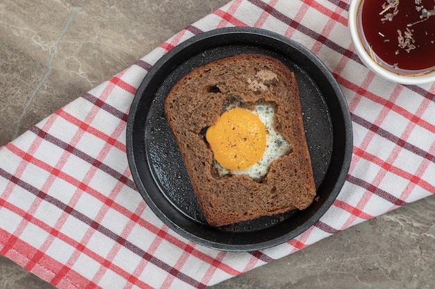 Жареное яйцо внутри ломтика черного хлеба на черной тарелке и чая. Фото высокого качества