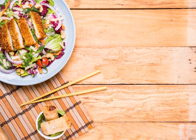 Foto gratuita insalata di filetto fritto con involtini primavera in ciotola di ceramica e bacchette contro tavolo in legno