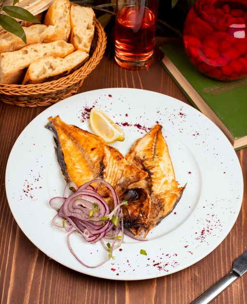 Жареная рыба на гриле подается в белой тарелке с луковым салатом, лимоном и зеленью