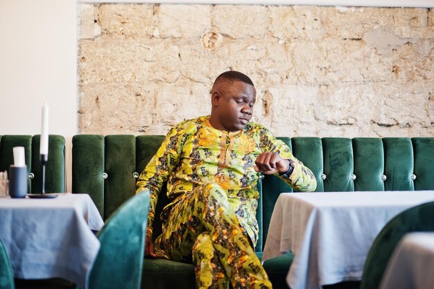 Friendly afro man in traditional yellow clothes resting on the couch at restaurant and looking at his watches on hand