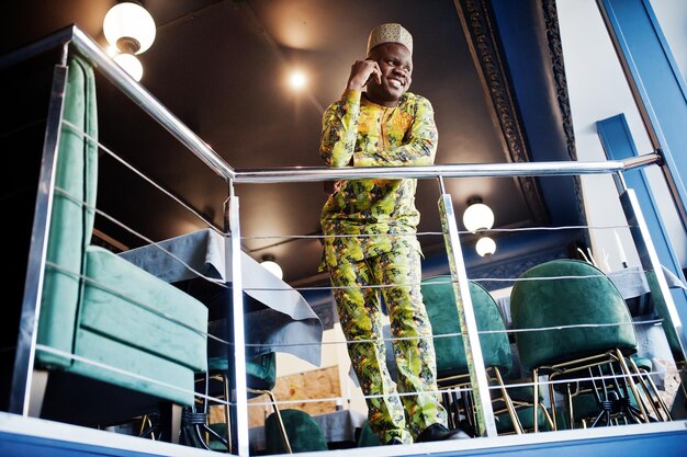 Friendly afro man in traditional yellow clothes with cap at restaurant near the handrail speaking at mobile phone