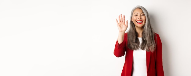 Friendly asian senior lady in elegant blazer saying hi waving hand and greeting you with happy smile