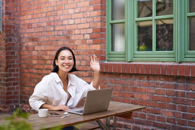 Friendly asian woman sitting with laptop waving at you saying hello hi gesture greeting you while wo