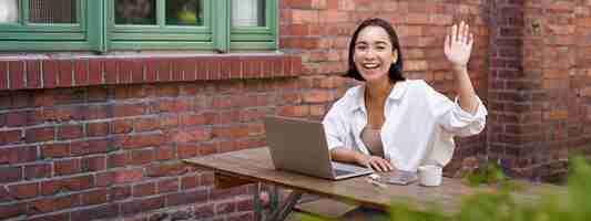 Free photo friendly asian woman sitting with laptop waving at you saying hello hi gesture greeting you while